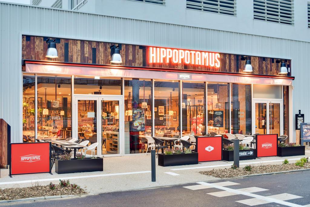 Extérieur moderne d'un restaurant Hippopotamus avec de grandes fenêtres. Terrasse extérieure et enseigne rouge vif sous les lumières du soir.