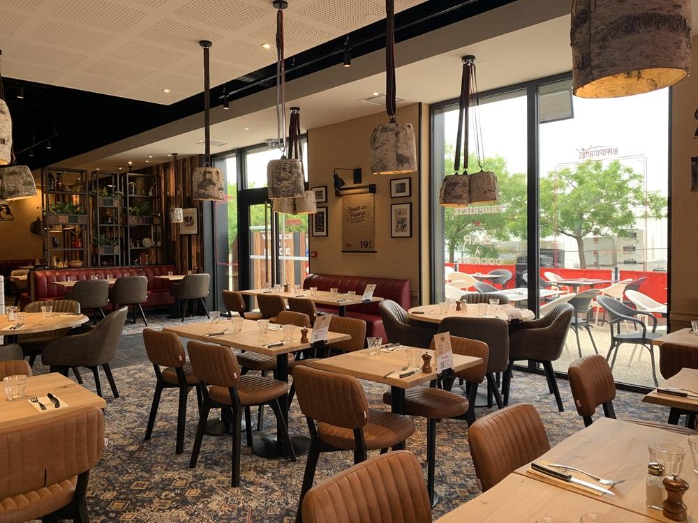 Intérieur d'un restaurant Hippopotamus chaleureux avec tables en bois, chaises rembourrées, suspensions lumineuses et grandes fenêtres donnant sur une terrasse avec des tables en plein air.