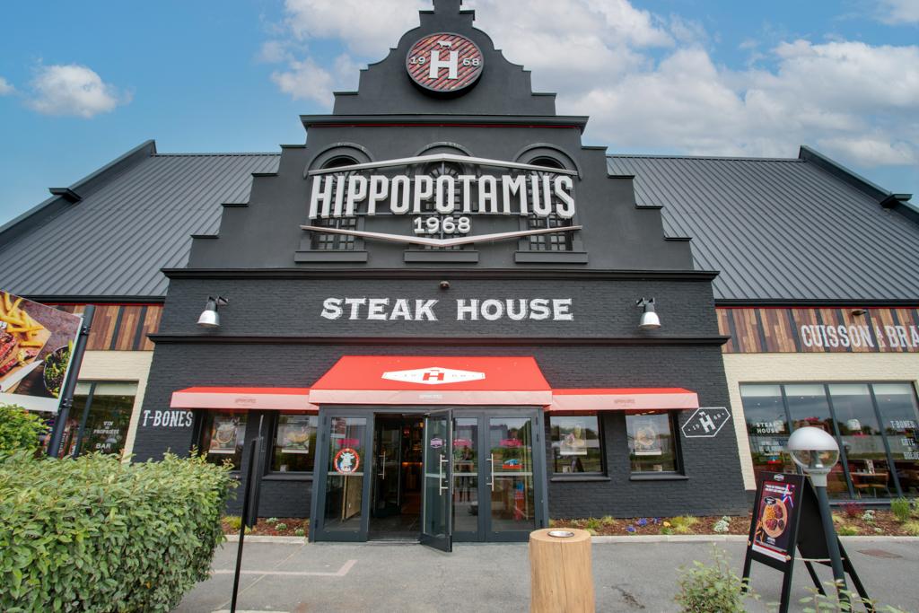 Extérieur du restaurant Hippopotamus Steak House avec son enseigne voyante, sa façade noire, son toit en pignon et son auvent rouge. Ciel bleu parsemé de nuages.