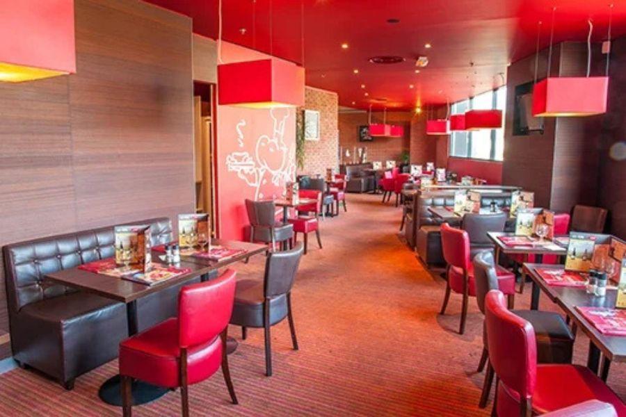 Intérieur chaleureux du restaurant avec une décoration rouge et marron, des lampes rouges au plafond, des tables marron foncé et des chaises rembourrées, créant une atmosphère chaleureuse et accueillante.