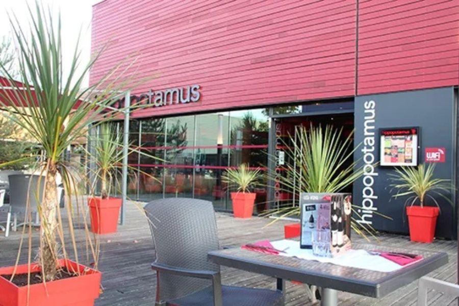 Un restaurant Hippopotamus moderne avec une façade rouge et de grandes plantes en pot sur une terrasse en bois. Une table est dressée avec un menu et des serviettes.