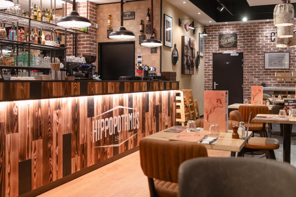Intérieur de restaurant Hippopotamus chaleureux avec comptoir en bois, sièges de bar et tables. L'éclairage tamisé et les murs en briques créent une atmosphère accueillante.