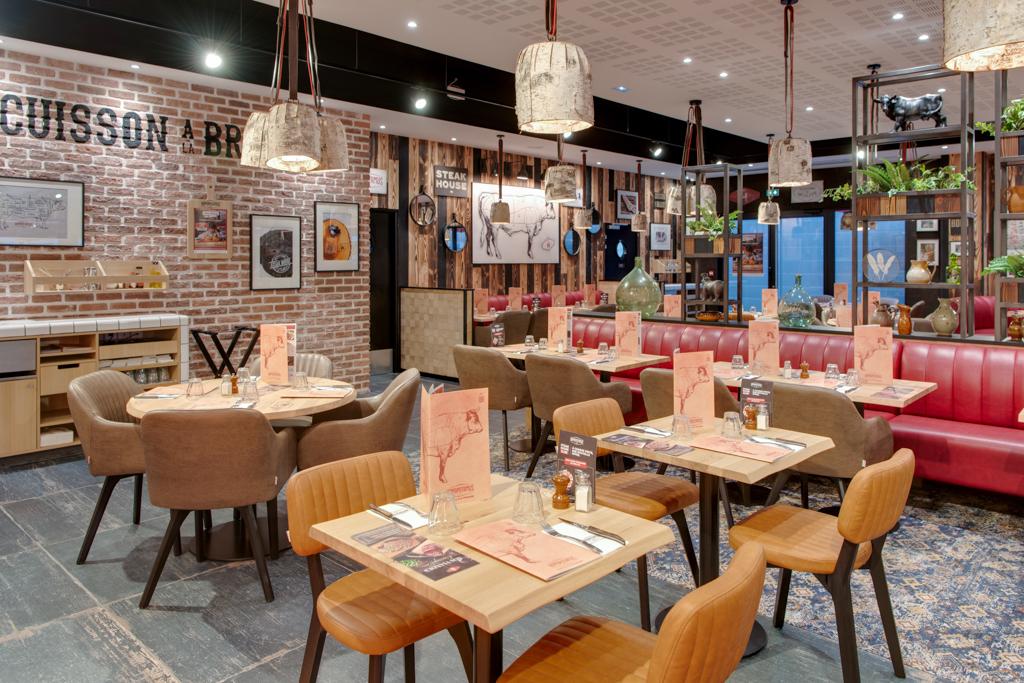 Un restaurant Hippopotamus à l'intérieur chaleureux avec des tables en bois, des banquettes rouges, des murs en briques, des suspensions lumineuses et une décoration éclectique.