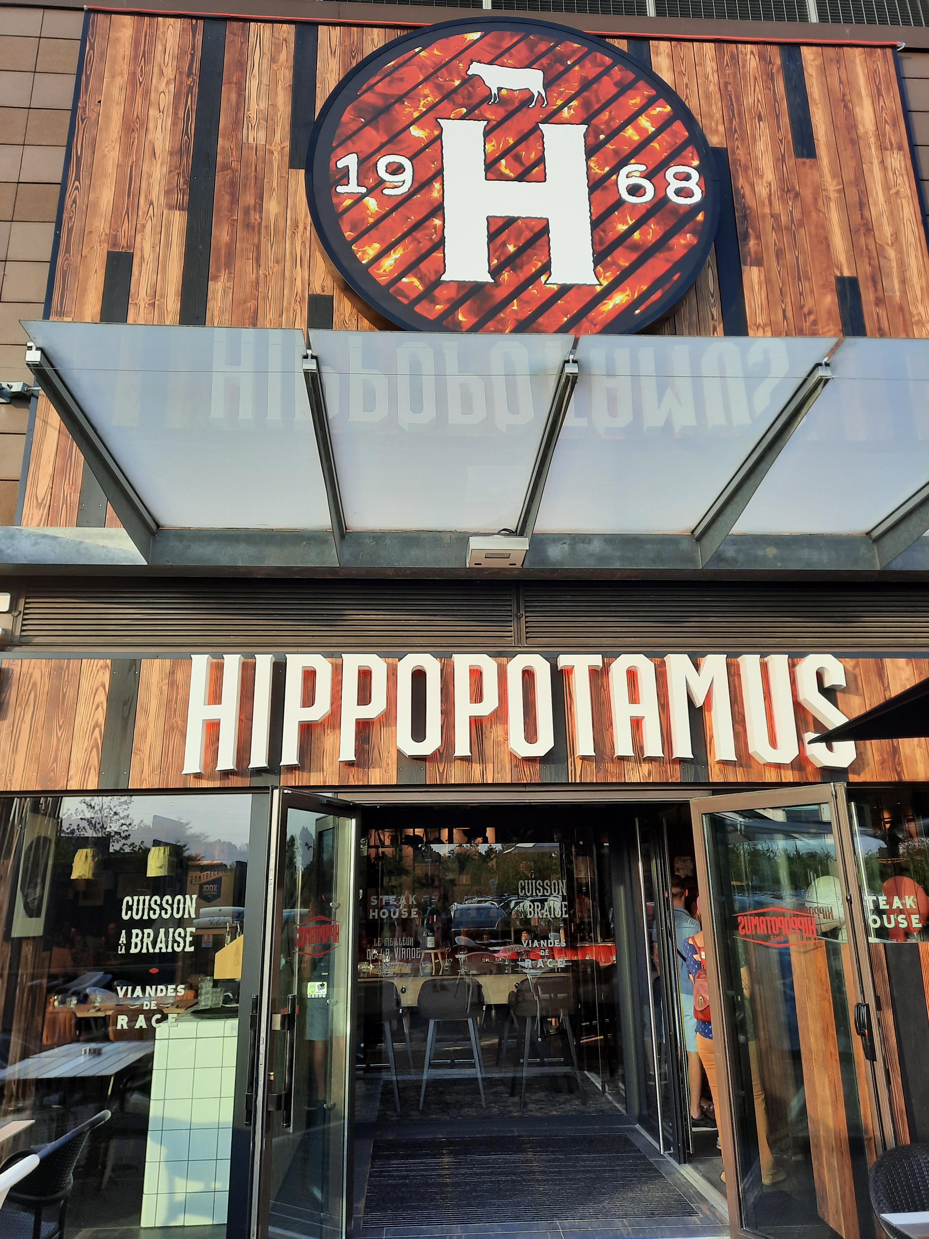Entrée d'un restaurant Hippopotamus avec décor en bois, grand « H » et logo 1968 au-dessus, et portes vitrées ouvertes menant à l'intérieur.