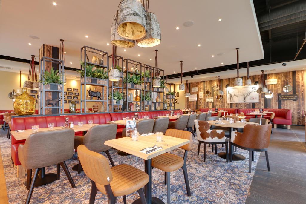 Intérieur de restaurant Hippopotamus moderne avec banquettes rouges, tables en bois, chaises assorties, suspensions lumineuses et étagères décoratives pour plantes. Touches rustiques partout.